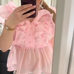 Rosa volangblus med knappar - Supersöt ljusrosa blus med volanger och knappar framtill. Blusen har korta ärmar och är tillverkad i ett lätt, transparent material som ger en luftig känsla. Perfekt för dig som gillar romantiska detaljer och en feminin stil.           Lånad bild!
