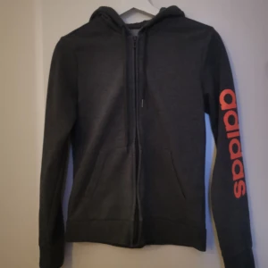 Mörkgrå zip-hoodie från Adidas - Mörkgrå hoodie med dragkedja från Adidas. Stor logga i rött längs ena ärmen och två fickor framtill. Perfekt för en sportig och avslappnad stil.