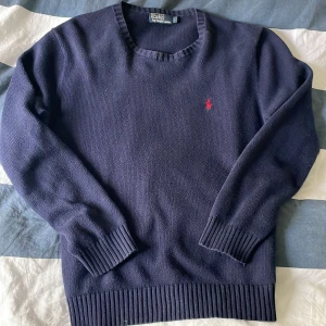 Mörkblå stickad tröja från Polo Ralph Lauren - Säljer en klassisk mörkblå stickad tröja från Polo Ralph Lauren med röd broderad logga på bröstet. Tröjan har rund halsringning och ribbade muddar vid ärmslut och nederkant. Perfekt för en stilren och tidlös look.
