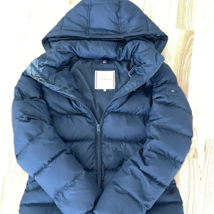 Mörkblå pufferjacka från Tommy Hilfiger - Säljer en mörkblå pufferjacka från Tommy Hilfiger med huva och dragkedja. Jackan är vadderad och har en klassisk, sportig look med diskret logga på ärmen. Perfekt för kyliga dagar och enkel att matcha med olika outfits. Strl 164, motsvarar ungefär xs. Skriv gärna vid ytterligare frågor!💘