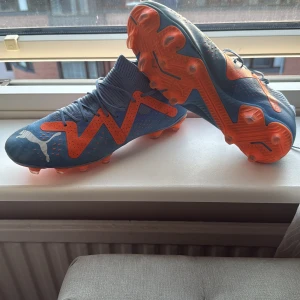 Blå och orange fotbollsskor från Puma - Säljer ett par blå fotbollsskor från Puma med orangea detaljer och snörning. Skorna har en stickad strumpliknande öppning och är designade för bra grepp på planen. Perfekta för dig som vill sticka ut på matchen! Använda kanske 5-6 gånger eftersom dom var för små 