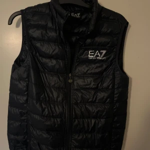 Svart dunväst från Emporio Armani EA7 - Snygg svart dunväst från Emporio Armani EA7 med quiltad design och dragkedja framtill. Västen har hög krage och logotyp på bröstet. Perfekt för lager-på-lager och stilrena outfits.