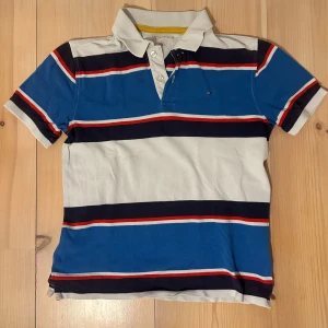 Randig pikétröja från Tommy Hilfiger - Säljer en klassisk pikétröja från Tommy Hilfiger med breda ränder i blått, vitt, svart och rött. Tröjan har korta ärmar, krage och knappar framtill. Perfekt för en avslappnad och stilren look.