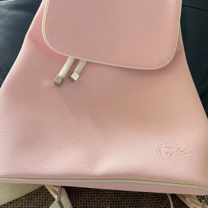 Lacoste damryggsäck  - Mycket fin och knappt använd rosa Lacoste damryggsäck med vita remmar. Mått c.a 32 cm höjd och 29 cm bredd. Pris 600kr plus porto.