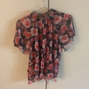 Blommig blus från Gina Tricot - Säljer en blommig blus från Gina Tricot med rosa och röda blommor på mörk botten. Blusen har korta ärmar och en högre rynkad halsringning. Perfekt för dig som vill ha en luftig och färgglad topp. genomskinlig 