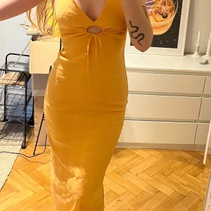 Aprikos långklänning med cut-out - Säljer en snygg aprikos långklänning med v-ringning och cut-out detalj framtill. Klänningen är ärmlös och har en slits bak. Perfekt för sommarens alla tillfällen! Materialet känns mjukt och följsamt, har lite stretch i sig 