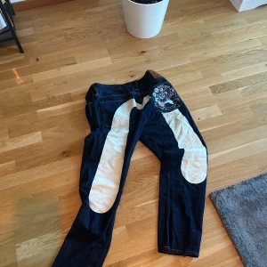 Mörkblå jeansbyxor från Evisu med broderad design design - Unika mörkblå jeans från Evisu med stora vita broderade detaljer på benen och en broderad Raijin-figur på baksidan. Klassisk läderpatch med Evisu-logga och japanska motiv. Perfekta för dig som vill sticka ut med din stil.