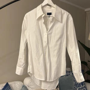 Vit skjorta från GANT - Klassisk vit skjorta från GANT med lång ärm och knappar framtill. Skjortan har en stilren design med bred krage och är tillverkad i bomull. Perfekt för en clean och tidlös look.