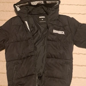 Svart pufferjacka från Hoodrich - Säljer en svart pufferjacka från Hoodrich med huva och dragkedja. Jackan har logga på bröstet och insidan av huvan. Perfekt för kyligare dagar och har en modern look.