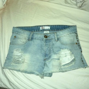 Ljusblå jeansshorts med fransar och slitningar - Säljer ett par ljusblå jeansshorts från Mossimo med fransig kant och slitna detaljer framtill. Shortsen har normal passform, klassiska fickor och nitar längs ena sidan. Perfekta för sommaren!