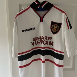 Manchester United retro fotbollströja - Klassisk vit Manchester United fotbollströja från Umbro med svart och röd detalj, klubbmärke på bröstet och sponsorlogga 'Sharp Viewcam' framtill. Tröjan har korta ärmar och svart-röda ränder över bröstet och magen.