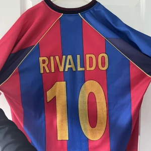 Barcelona fotbollströja Rivaldo 10  - Klassisk Barcelona fotbollströja med Rivaldo och nummer 10 på ryggen. Tröjan är randig i rött och blått med guldiga detaljer och klubbmärke på bröstet. Kortärmad modell i lätt och ventilerande material.