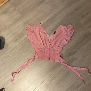 Rosa prickig omlottopp från SHEIN - Supersöt rosa omlottopp från SHEIN med vita prickar. Toppen har volangdetaljer vid axlarna, smockad midja och knytband i sidan. Perfekt för vår och sommar när du vill ha något lätt och luftigt.