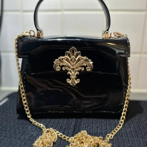 Svart handväska med gulddetaljer och kedja - Säljer en svart, glansig handväska med guldfärgad kedja och dekorativt guldfäste framtill. Väskan har ett handtag upptill och avtagbar axelrem i kedja. Perfekt för dig som vill ha en elegant accessoar med lyxig känsla.