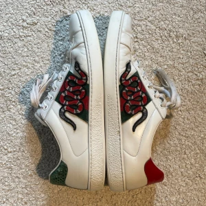 gucci ace snake - Säljer ett par vita Gucci Ace sneakers i skinn med ikonisk röd och grön orm brodyr på ytter sidan. Skorna har gröna och röda detaljer på hälen samt klassiska Gucci-ränder på inner sidan. Perfekta för dig som vill sticka ut med lyxig streetstyle.