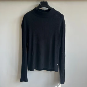 Our Legacy Artist Turtleneck svart - Svart mockneck tröja från Our Legacy med stickad struktur och hög hals. Tröjan har en avslappnad passform och diskreta slitningar för en edgy look. Perfekt för dig som gillar minimalistisk stil med en twist. 