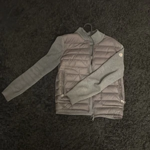 Grå cardigan jacka från Moncler - Snygg grå cardigan jacka från Moncler med quiltad front och mjuka stickade ärmar. Jackan har dragkedja framtill och två fickor med dragkedja. Perfekt för lager-på-lager och stilren look.