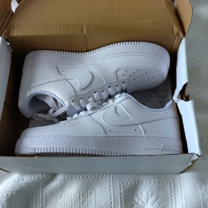 Nike Air Force 1 vita sneakers - Klassiska helvita Nike Air Force 1 sneakers med låg profil. Skorna har rund tå, platt sula och är tillverkade i skinn med perforerade detaljer på tån. Perfekta för dig som gillar en clean och tidlös stil.