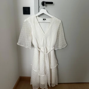 Klänning från Vero Moda - Superfin vit klänning från Vero Moda med diskreta prickar, v-ringning och volangdetaljer. Klänningen har korta, transparenta ärmar och ett knytband i midjan. Perfekt för sommarens tillfällen!