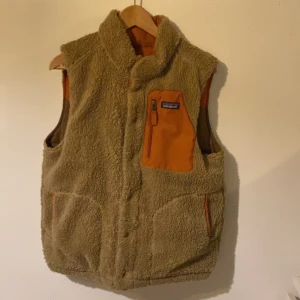 Beige fleeceväst från Patagonia - Säljer en beige fleeceväst från Patagonia med orange detaljer och bröstficka med dragkedja. Västen har hög krage och två sidofickor med orange kant. Perfekt för lager-på-lager under kyliga dagar.
