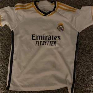 Real Madrid fotbollströja - Säljer en vit Real Madrid fotbollströja från Adidas med gula och mörkblå detaljer. Tröjan har klubbmärket på bröstet och sponsortryck 'Emirates Fly Better' på framsidan. Klassisk design med korta ärmar och rund hals. Lite fläckar på tröjan 