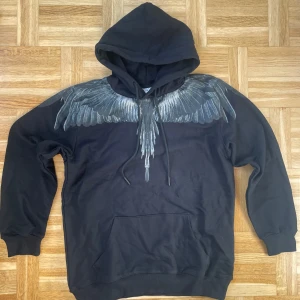 Svart hoodie  från Marcelo Burlon - Säljer en svart hoodie från Marcelo Burlon County of Milan med ikoniskt vingmönster i grått och vitt över axlar och bröst. Tröjan har huva med dragsko och en stor magficka framtill. Perfekt statement-plagg för streetwear.