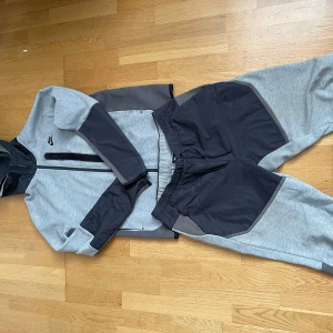 Nike tech hybrid tracksuit/mjukisdress - Säljer ett par grå mjukisbyxor från Nike med svarta detaljer och dragkedjeficka. Byxorna har elastisk midja och är perfekta för en avslappnad stil. Materialet är mjukt och bekvämt, och byxorna har en sportig look.