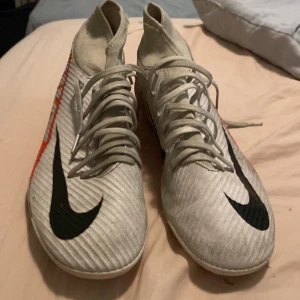 Nike mercurial clubs - Säljer ett par vita Nike Mercurial fotbollsskor med svarta och röda detaljer. Skorna har snörning och en högre strumpliknande krage för extra stöd. Perfekta för dig som vill ha stilrena och snabba skor på planen.