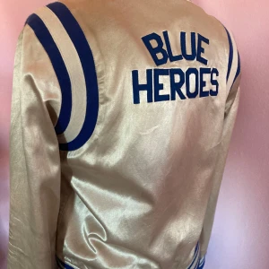 Silver/dustfärgad bomberjacka med text - Snygg bomberjacka i silver med blå detaljer och texten 'BLUE HEROES' på ryggen. från Maison Scotch. Jackan har blå ränder på axlarna, ribbade muddar och knappar framtill. Perfekt för dig som vill sticka ut med en retrostil.