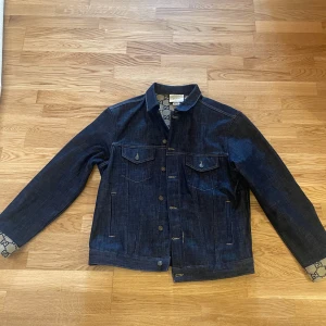 Mörkblå jeansjacka från Gucci - Säljer en mörkblå jeansjacka från Gucci med ikoniskt GG-mönstrat innerfoder och detaljer på ärmslut. Klassisk modell med knappar framtill och två bröstfickor. Perfekt för dig som vill ha en lyxig och stilren jacka med unika detaljer.