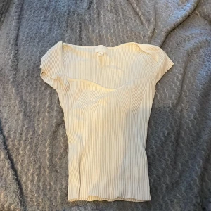 Beige ribbad topp från H&M - Säljer en beige ribbad topp från H&M i storlek XS. Toppen har korta ärmar och en figurnära passform med snygga sömdetaljer framtill. Perfekt att styla till olika outfits!