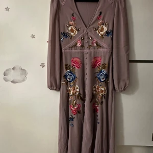 Långklänning med broderade blommor från Hope & Ivy - Superfin långklänning i dov lila nyans från Hope & Ivy. Klänningen har v-ringning, långa ärmar och vackra broderade blommor i rosa, blått och beige framtill. Perfekt för dig som vill sticka ut med detaljerad brodyr och romantisk känsla.