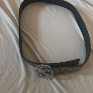 Gucci bälte med silverfärgat spänne - Säljer ett Gucci bälte med klassiskt GG-mönster och stort silverfärgat GG-spänne. Bältet är brett och har beige och grå färgtoner med tydligt Gucci-mönster. Perfekt accessoar för att lyfta din outfit.