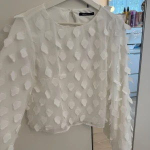Vit blus med 3D-detaljer från Gina Tricot - Superfin vit blus från Gina Tricot. Blusen har lång ärm och rund halsringning, samt en liten knappöppning bak i nacken. Perfekt för dig som vill sticka ut lite extra!