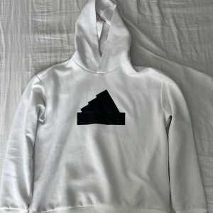 Vit hoodie från Adidas - Säljer en stilren vit hoodie från Adidas med svart logga framtill. Tröjan har långa ärmar, ribbade muddar och huva. Perfekt för en sportig och avslappnad look.