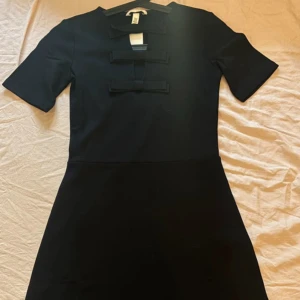 Svart klänning med rosetter från HM - Snygg svart klänning från HM med kort ärm och tre dekorativa rosetter framtill. Klänningen har en klassisk, figurnära passform och är perfekt för dig som gillar stilrena detaljer.