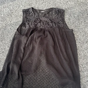 Svart ärmlös blus med spets från KappAhl - Svart ärmlös blus från KappAhl med vacker spets upptill och lätt transparent tyg nedtill. Blommigt spetsmönster över bysten och mjukt fallande kjol. Perfekt för dig som gillar romantiska detaljer. Det är tunt och genomskinligt typ så de passar jättebra med ett svart linne under