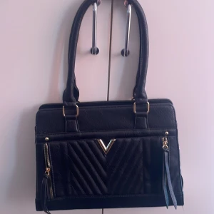 Svart handväska med gulddetaljer - Säljer en svart handväska med två rejäla handtag och guldfärgade detaljer. Väskan har ett V-format mönster framtill, dragkedjor på sidorna och dekorativa tofsar. Perfekt storlek för vardagens alla prylar. Prislapp kvar!