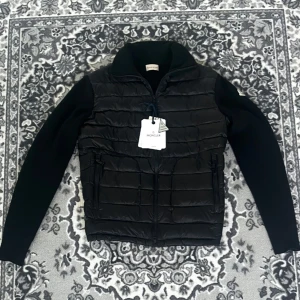 Svart dunväst med ärmar  - Snygg svart dunväst med långa stickade ärmar och quiltad framsida. Västen har dragkedja framtill och två fickor med dragkedja. L/XL