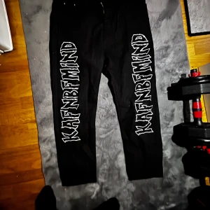 Svarta byxor med vit text från KAFFNBFMIND - Svarta byxor med raka ben och stor vit text 'KAFFNBFMIND' tryckt längs båda benen. Byxorna har en enkel design utan synliga detaljer som dragkedja eller knappar framtill. Perfekta för en streetwear-look.