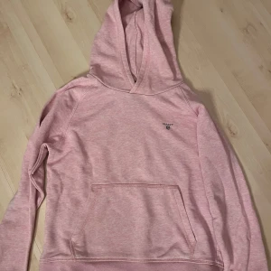 Ljusrosa hoodie från GANT - Säljer en ljusrosa hoodie från GANT med klassisk känguruficka framtill och liten logga på bröstet. Perfekt för en avslappnad stil och supermjuk att ha på sig. Passar dig som gillar bekväma och snygga tröjor!