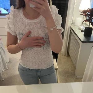 Vit spetsblus med volang från Zara - Säljer en söt vit spetsblus från Zara med korta ärmar och volangdetaljer på axlarna. Blusen har ett fint spetsmönster och rund halsringning. Perfekt för vår och sommar!