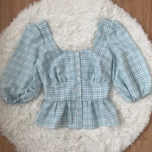 Blåvit rutig blus från New Look - Supersöt blus i blåvitt rutigt från New Look. Modellen har puffiga långärmar, knappar framtill och volang nertill. Passar storlek 32-34.