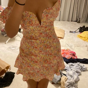 Blommig kortklänning med volang - Supersöt kortklänning med blommigt mönster i rosa, gul och vit. Klänningen har en v-ringad offshoulder-överdel och volangkant nertill. Perfekt för sommarens alla tillfällen!