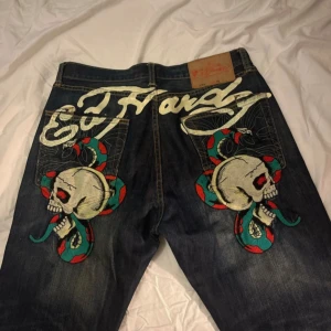 Ed Hardy jeans med broderade dödskallar och ormar - Säljer ett par mörkblå jeans från Ed Hardy med stora broderier av dödskallar och ormar på bakfickorna. Snyggt Ed Hardy-tryck både fram och bak. Klassisk femficksmodell i bomull med raka ben och normal passform. Jeansen är även från LOT 2009 begränsad upplaga i ofantligt bra skick. Små skador vid hälar förekommer! Tveka inte om fler frågor!