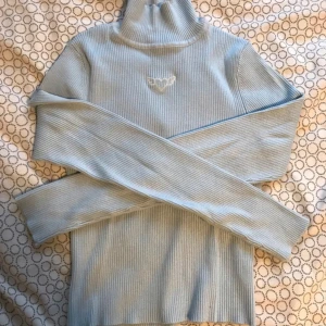 Ljusblå ribbad polotröja från Divided - Säljer en ljusblå ribbad polotröja från Divided. Tröjan har lång ärm, hög krage och ett litet broderat fjärilsmotiv framtill. Perfekt för lager-på-lager och enkel att matcha med jeans eller kjol.