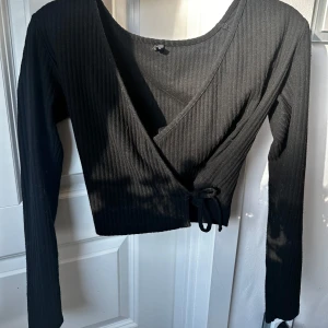 Svart omlottopp från ONLY - Säljer en svart ribbad omlottopp från ONLY i storlek XS. Toppen har långa ärmar, croppad passform och knyt i sidan. Perfekt för dig som gillar stilrena och enkla plagg med en twist. Använt många gånger men är i bra skick ❤️