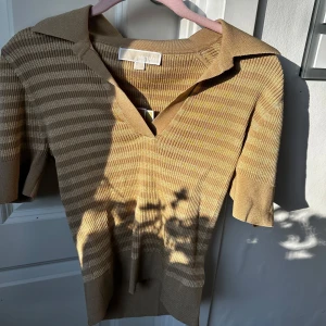Beige stickad pikétröja från Michael Kors - Säljer en beige stickad pikétröja från Michael Kors med smala horisontella ränder och v-ringning. Tröjan har korta ärmar och klassisk krage. Perfekt för en stilren och avslappnad look. Helt ny säljer då den inte riktigt är min stil! ❤️