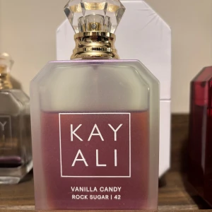 Kayali Vanilla Candy Rock Sugar 42 EdP - En söt och gourmandig parfym med noter av kanderade päron, bubbelgummi och stensocker. Flaskan är matt lila med guldigt lock och tydlig vit logotyp. Doften är varm, kryddig och har inslag av vanilj, tonkaböna, sandelträ och patchouli.