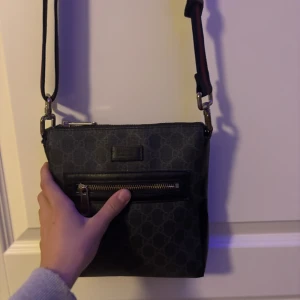 Svart axelväska med mönster och dragkedja - Säljer en svart axelväska från Gucci med diskret mönster och justerbar axelrem med röd och grön rand. Väskan har en praktisk ytterficka med dragkedja och är perfekt för att bära det viktigaste. Snygg och stilren design.